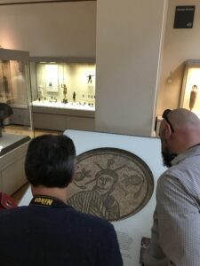 In a London museum gallery, during a Christian tour, two people examine a mosaic artwork. The mosaic depicts a robed figure with a raised hand, surrounded by intricate patterns. Display cases with artifacts are visible in the background.