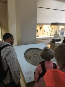 People on a Christian Tour are observing a mosaic displayed in a London museum exhibit. Several art pieces sit in the background under glass cases, while the light-colored surrounding walls add brightness to the space.
