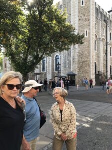 A group of people stands in a sunlit courtyard, possibly part of an ancient castle in London. A large tree offers shade, while others from the Christian tour explore the historic site, adding life to the scene with their presence near the old stone building.