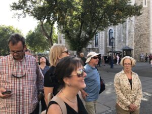 A group of people stands outside a historic stone building, possibly on a Christian tour in London, with trees in the background. One person checks their phone while others, sporting sunglasses, enjoy the sunny and pleasant scene.