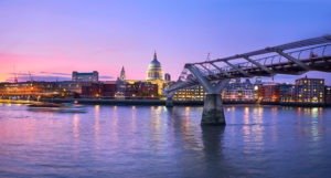 View of London at dusk with the Millennium Bridge crossing the Thames. As part of a London Christian Tour, St. Pauls Cathedral stands illuminated in the background against a colorful sky. A blurred boat is seen on the river, reflecting the city lights on the water.