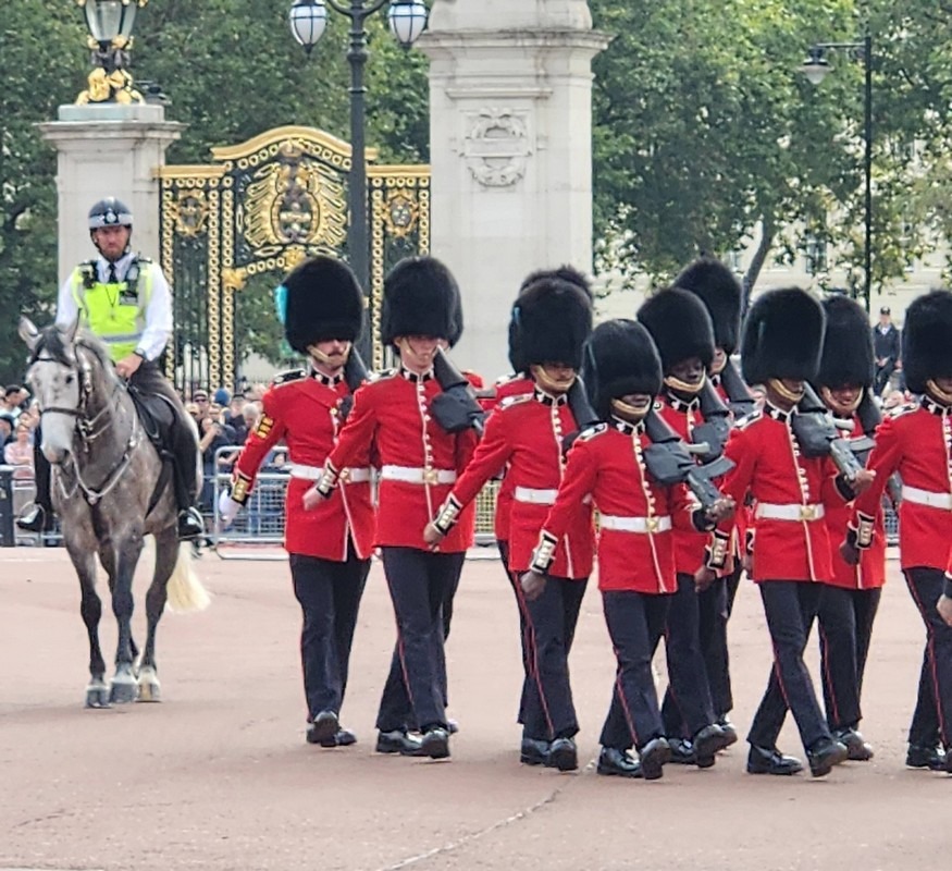 A group of ceremonial guards in red uniforms and tall black hats march in formation near an ornate gate, a classic sight during the London Christian Tour. A police officer on a horse oversees the event, set against a backdrop of lush green trees and lamp posts.
