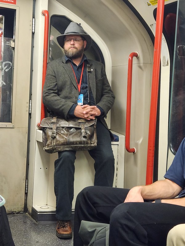 Nate Loper sits on a folding seat in the London subway. Hes carrying a large leather bag and has a red lanyard with a badge reading Christian Tour around his neck. Other passengers are partially visible.