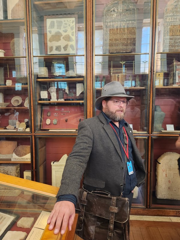 Nate Loper stands in a museum, leaning on a display case during the London Christian Tour. Behind him are glass cabinets filled with stone artifacts and inscriptions. He's dressed in a blazer and carries a leather satchel.