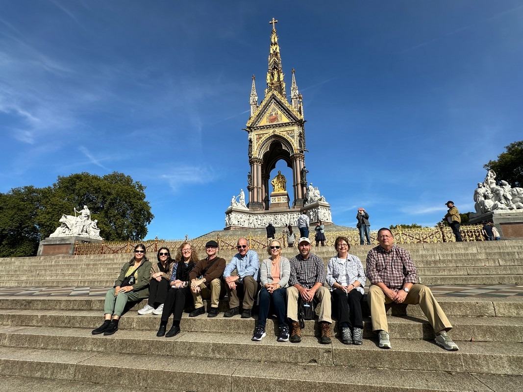 A group of people sits on the steps in front of the Albert Memorial during a sunny day on their London Christian Tour. The ornate monument, with its golden statue and intricate sculptures, is surrounded by green trees and clear blue skies.