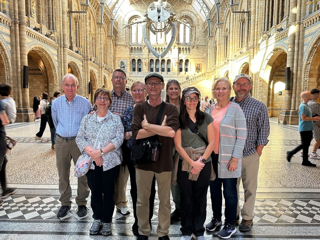 A group of nine people stands together inside a large, ornate hall with high arched ceilings and intricate architectural details, reminiscent of a classic London Christian tour. They face the camera, two donning hats, as bright lighting highlights the halls patterned floor.