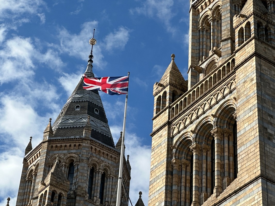 The image captures a close-up of the Natural History Museum in London, showcasing its intricate stone architecture. A British flag waves proudly on a pole, set against a blue sky with scattered clouds. A highlight on any London Christian tour, this landmark reflects rich cultural heritage.
