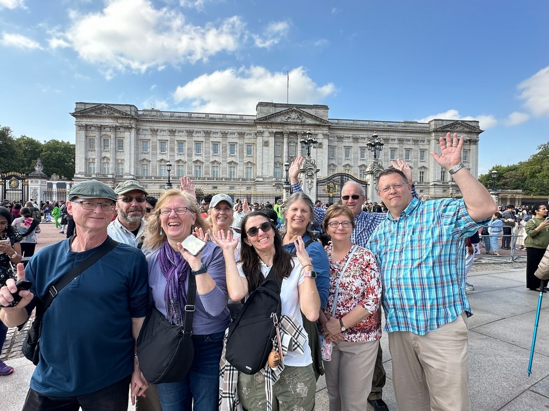 A group of people smiling and waving in front of Buckingham Palace on a sunny day are part of a lively London Christian Tour. They stand amidst the crowd, some holding bags and cameras, as the clear blue sky enhances their joyful experience.