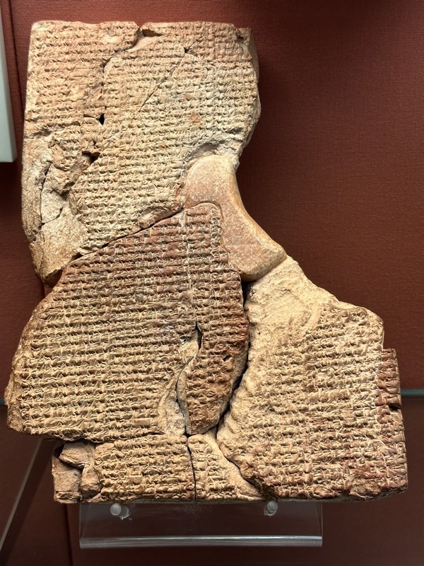 An ancient clay tablet with cuneiform script is displayed on a stand against a brown background, embodying age and historical significance. Like an artifact featured in a London Christian Tour, this fragmented piece resonates with stories of the past etched into its weathered surface.
