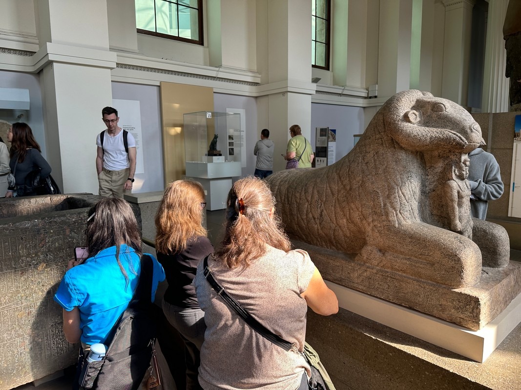 Visitors on the London Christian Tour admire an ancient stone statue of a reclining ram in the museum. The well-lit room invites reflection, with people exploring various exhibits. The atmosphere remains calm and educational, enriching their historical journey.