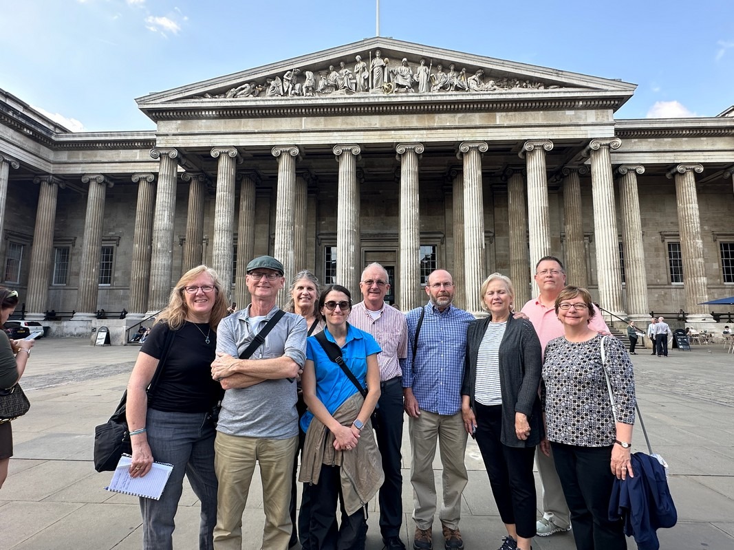 A group of nine people stands smiling in front of the British Museum with large columns and a pediment. The sky is clear, and the group appears to be enjoying their London Christian tour on this beautiful day out.