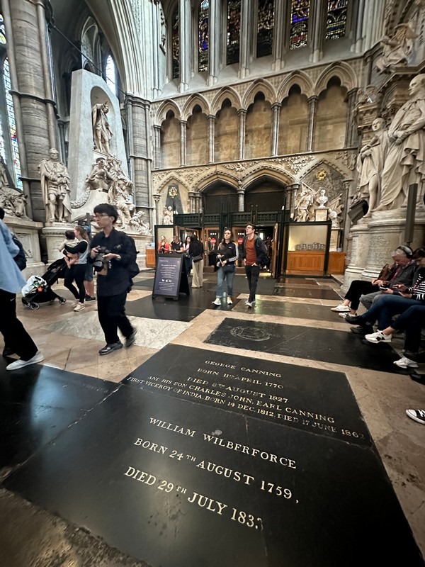 Interior of a cathedral with a stone floor displaying engraved memorials, including one for William Wilberforce. Visitors on the London Christian Tour walk through, observing the architecture and sculptures. Stained glass windows add a colorful backdrop to this spiritual journey.