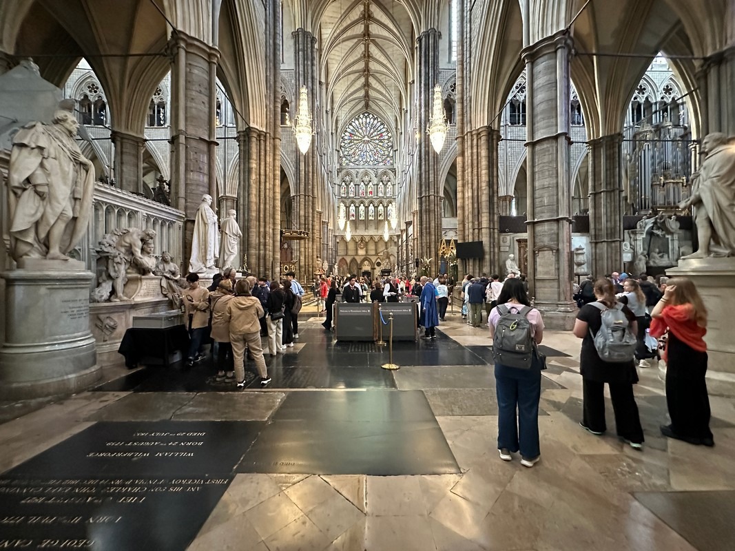 A large group of people stands inside a grand cathedral with high arched ceilings, part of the fascinating London Christian Tour. Ornate statues and a stained glass window are visible. The interior is spacious and historical, with the crowd gathered around a central area.