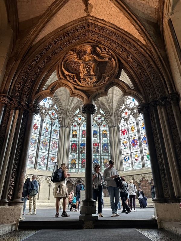 People on a London Christian Tour walk inside a historic building with ornate Gothic architecture. They are framed by an arched doorway with intricate carvings. Large stained glass windows in the background allow natural light to stream into the space.