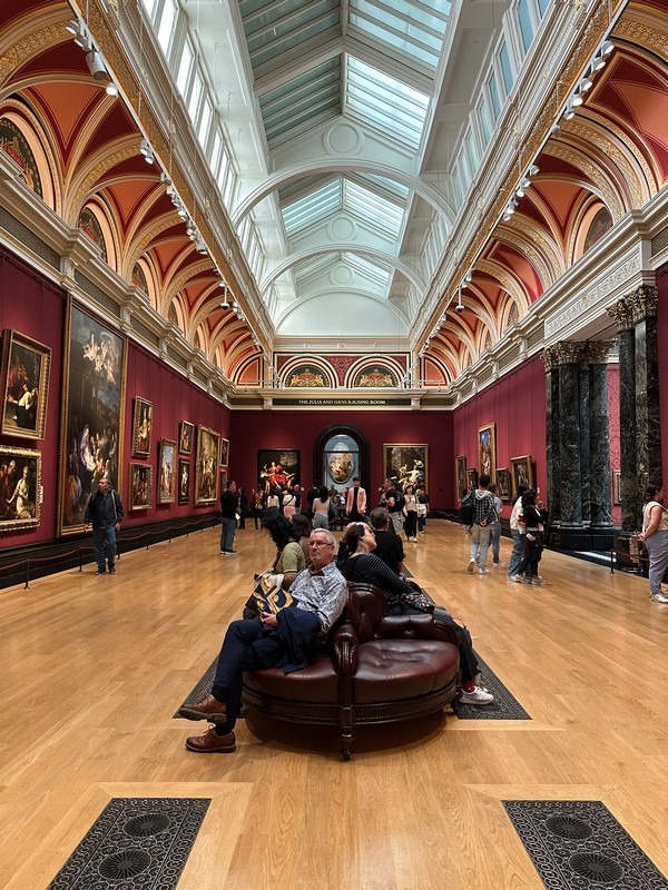 A spacious art gallery with high ceilings and skylights, featuring red walls adorned with classical paintings, is a highlight of the London Christian Tour. Visitors observe the artwork, and a few people sit on a central round leather bench.