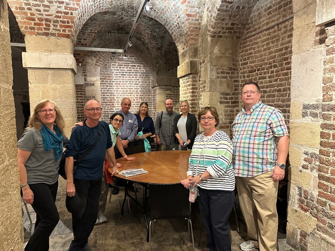 A group of nine people stands around a large wooden table in a room with brick walls and arched ceilings, reminiscent of historic London charm. They are casually dressed, smiling, and gathering as part of their enriching London Christian tour experience.