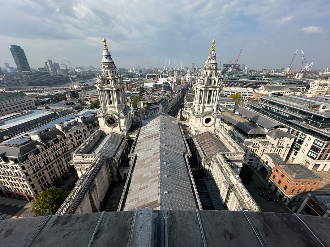 Aerial view of St. Pauls Cathedral in London, a highlight of the London Christian Tour, showcasing its twin towers and intricate architecture. The cityscape stretches into the distance under a partly cloudy sky, with cranes visible on the horizon.