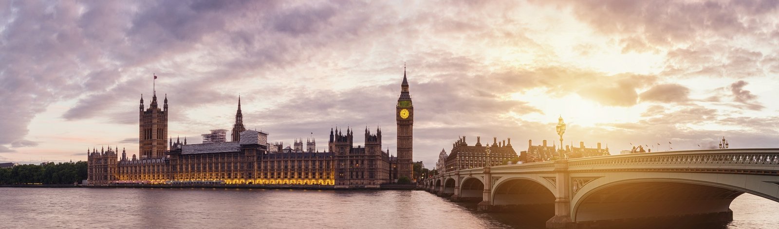 Panoramic view of the Houses of Parliament and Big Ben in London at sunset, with the River Thames in the foreground and Westminster Bridge to the right. The sky is partly cloudy with a warm glow from the setting sun.