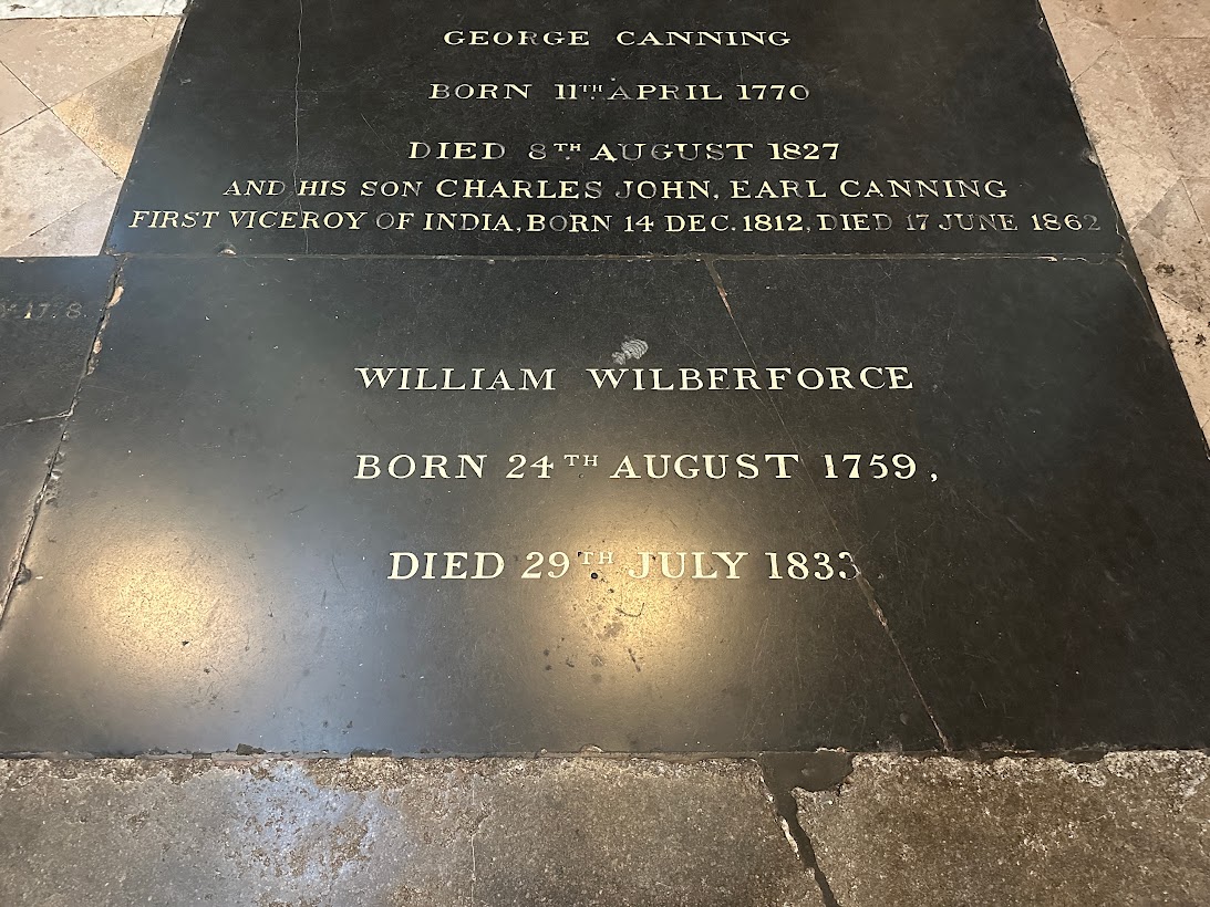 A stone memorial plaque engraved with the names and dates of George Canning, Charles John Earl Canning, and William Wilberforce, noting their birth and death dates. The text is etched in capital letters on dark stone.