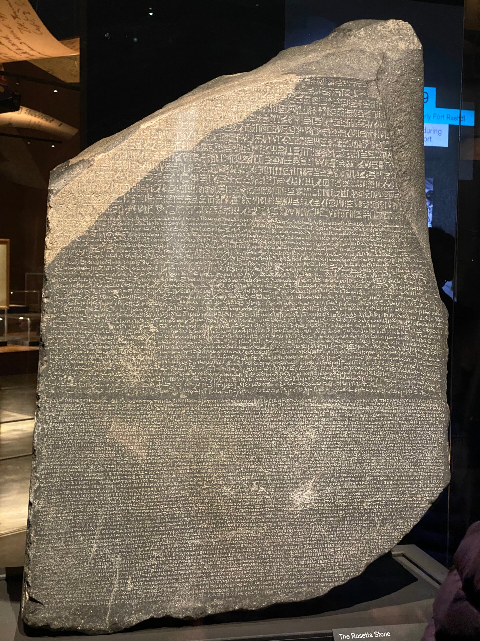 The Rosetta Stone, a large dark stone slab with inscriptions in three scripts—hieroglyphic, Demotic, and Ancient Greek—is displayed upright under glass in a renowned Christian London museum exhibit.