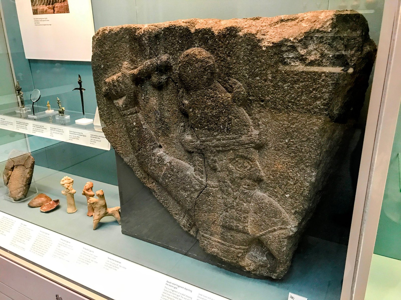 Hittite Culture in the British Museum, Nate Loper. A stone relief depicting a figure wearing a headdress and holding a weapon, displayed in a museum case with other ancient artifacts and descriptive labels. London Christian Tours