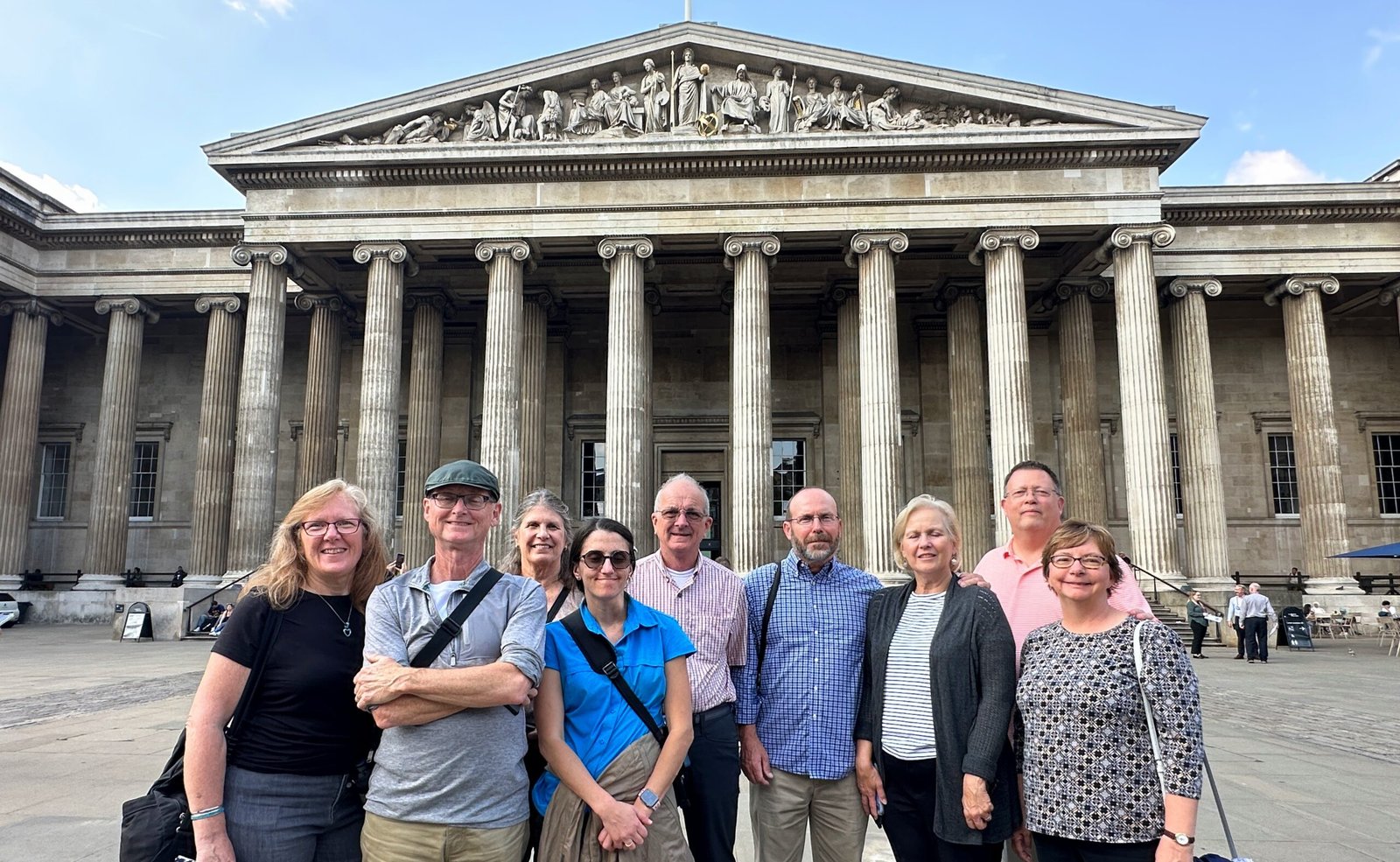 A group of nine adults stands smiling in front of the British Museum’s grand entrance with tall columns and classical pediment on a sunny day.