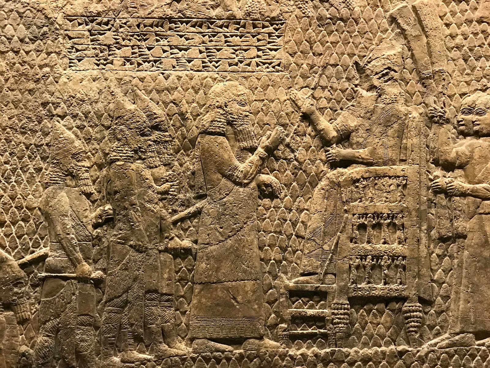 Assyrian Siege of Lachish Sennacherib British Museum Nate Loper. Ancient stone relief depicting figures in elaborate robes; one seated on a throne with others standing before, gesturing as if in conversation. The background features intricate carved patterns. London Christian Tours