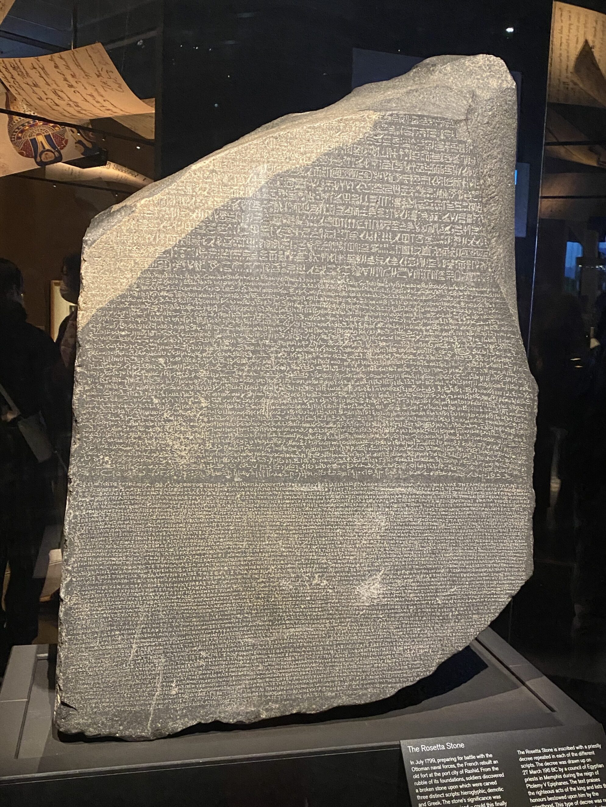 The Rosetta Stone, a large dark stone slab with inscriptions in three scripts, is displayed upright in a museum setting under focused lighting.