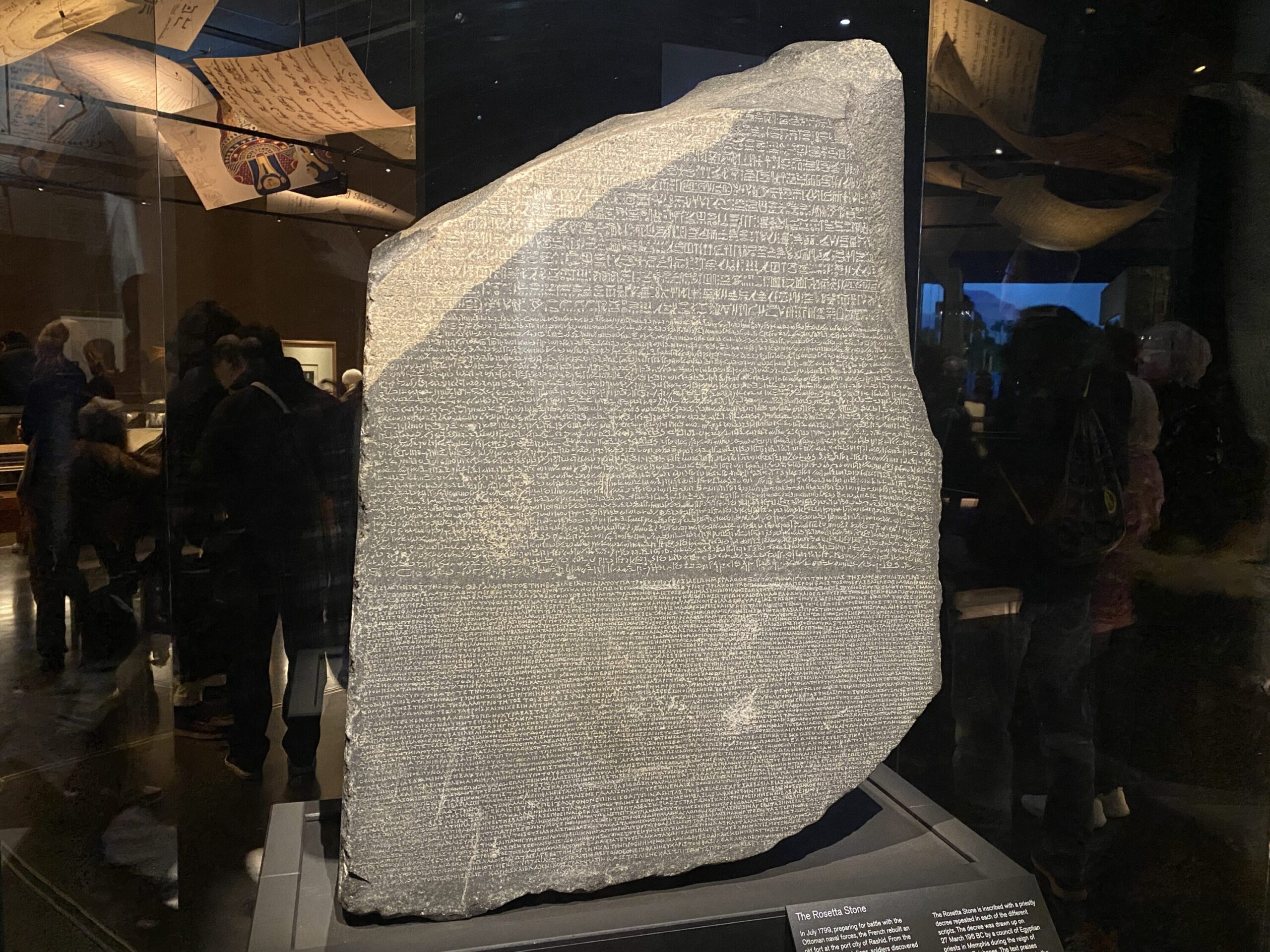 A large, inscribed stone slab (the Rosetta Stone) displayed in a glass case at a museum, surrounded by dim lighting and visitors in the background.