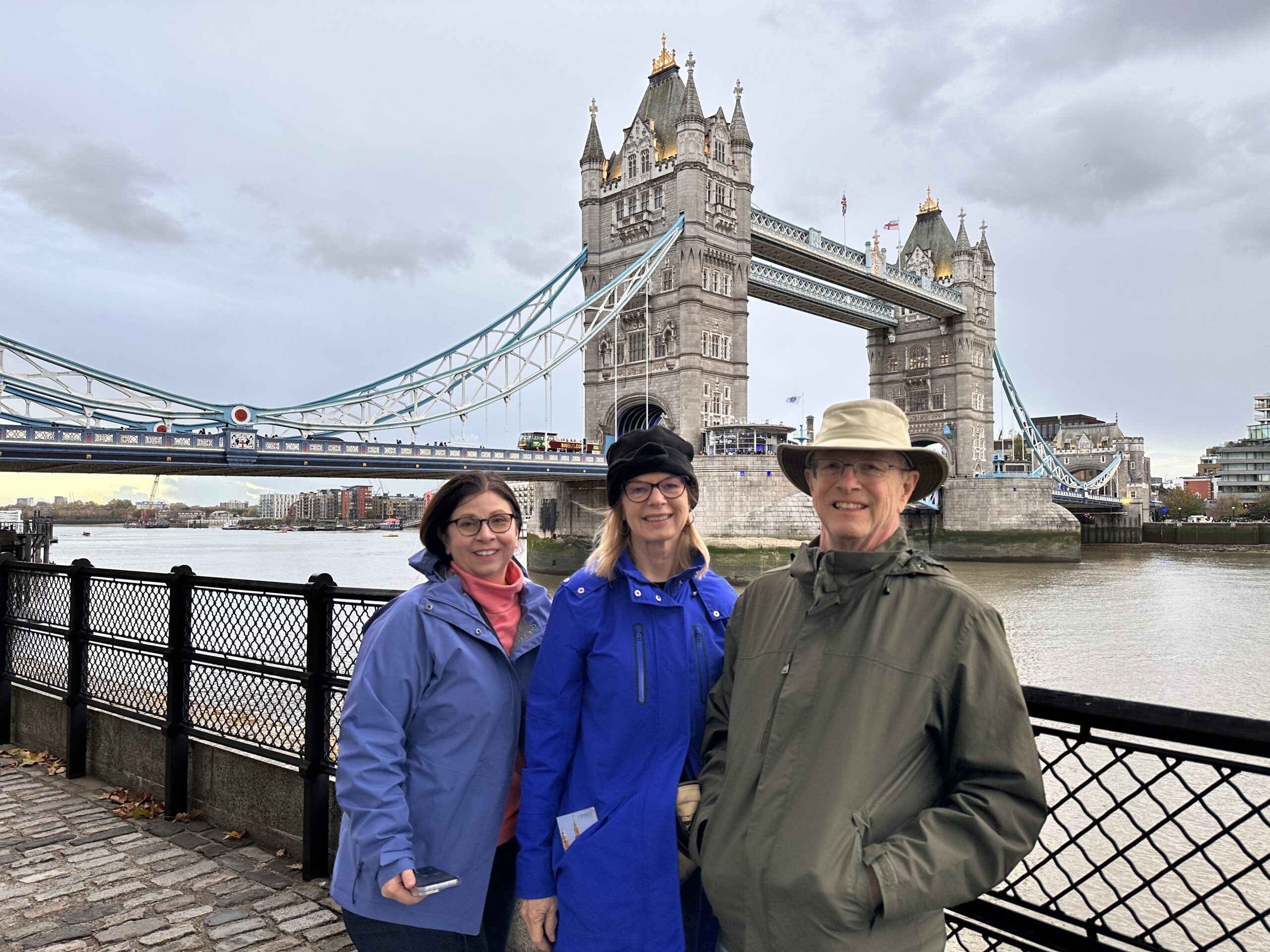 Three people in jackets stand smiling by the Thames River with Londons Tower Bridge in the background on a cloudy day.