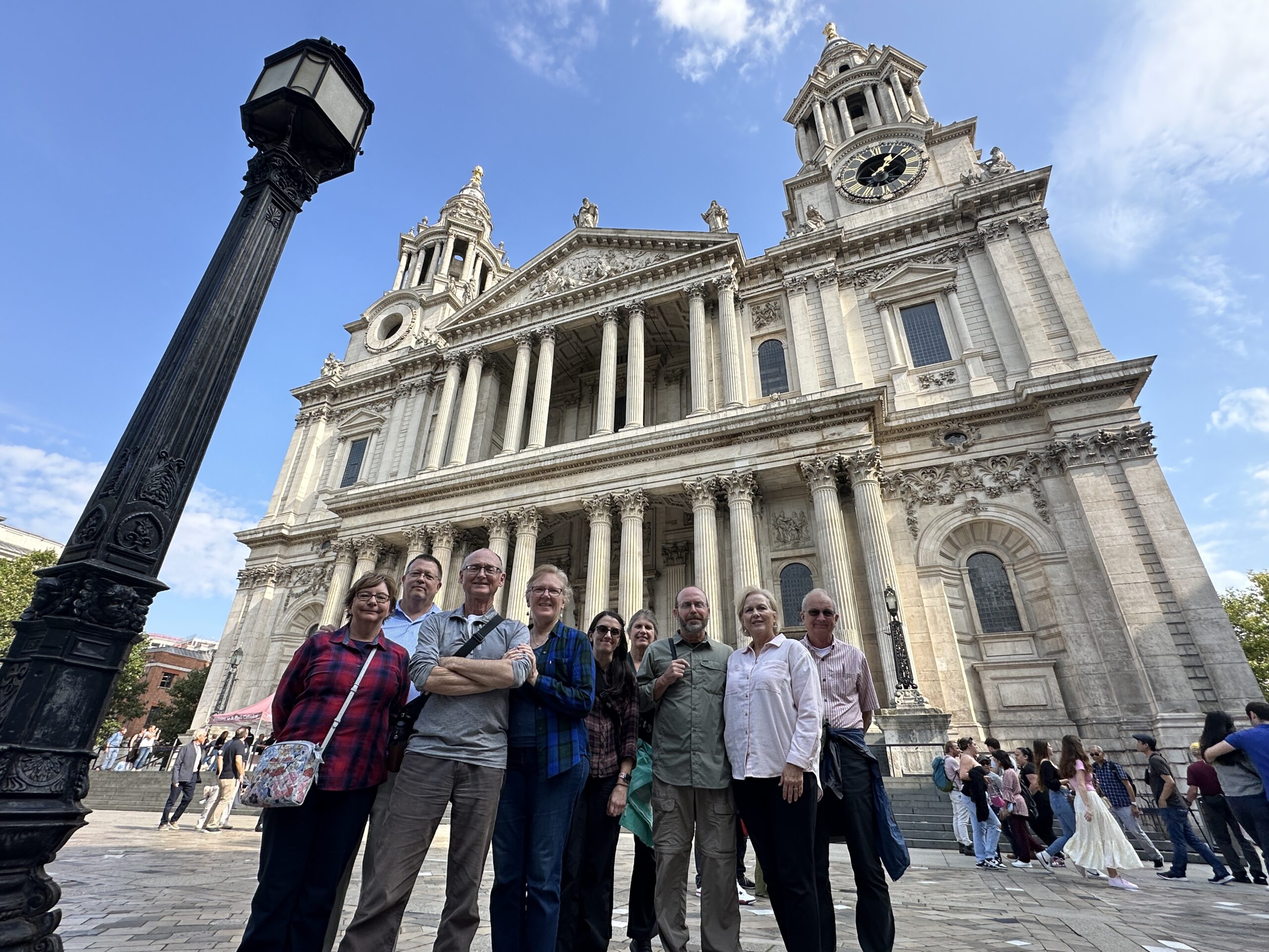 A group of nine people stand smiling in front of St. Pauls Cathedral in London on a sunny day, with the cathedrals grand facade and clock visible behind them.