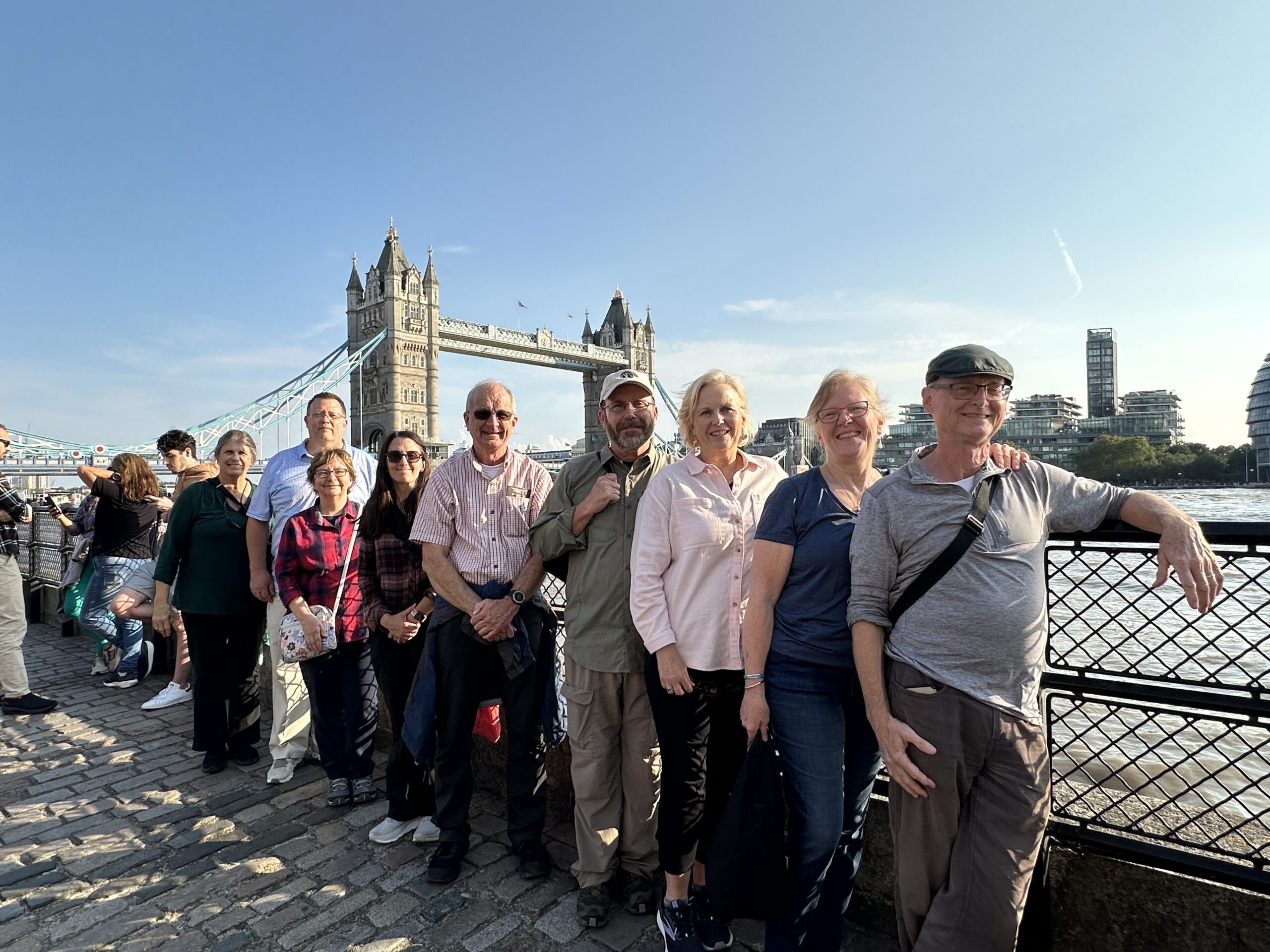 A group of people stands in a line smiling by the river with Londons Tower Bridge in the background on a sunny day.