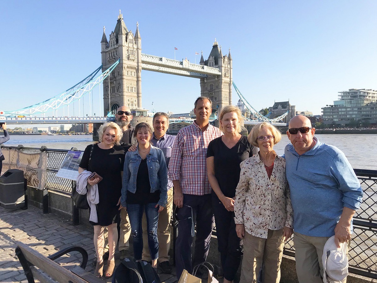 A group of eight adults poses and smiles in front of Tower Bridge in London on a sunny day by the Thames River.